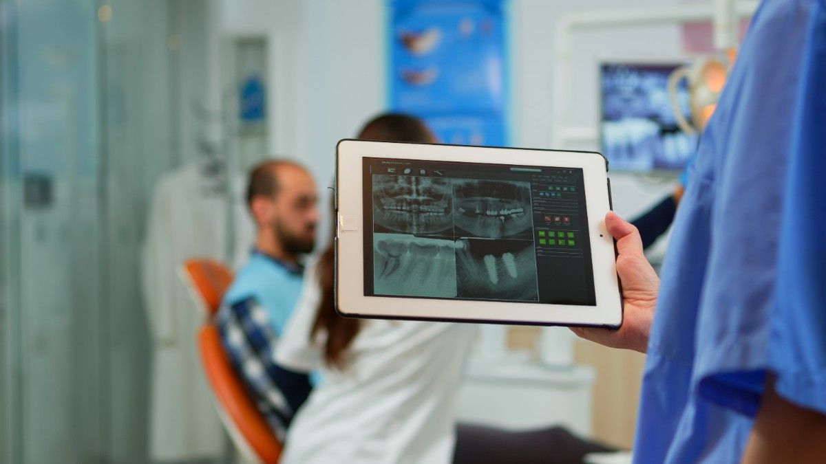 Dentist holding tablet displaying dental X-rays with patient in background